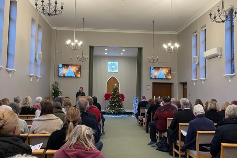 Photo of the inside of the crematorium, people are sitting in rows of chairs.