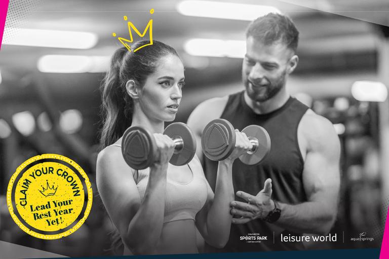 A man and a woman in a gym. The woman is using dumbbells while the main looks on encouragingly.h