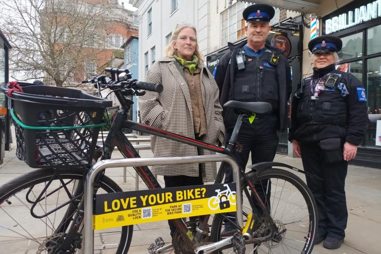Three people stand beside a bicycle secured to a bike rack on Colchester High Street with a sign promoting secure bike parking.