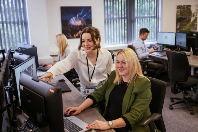 Someone sitting at a desk in front of a computer smiling with someone standing next to them pointing at the screen on the computer smiling