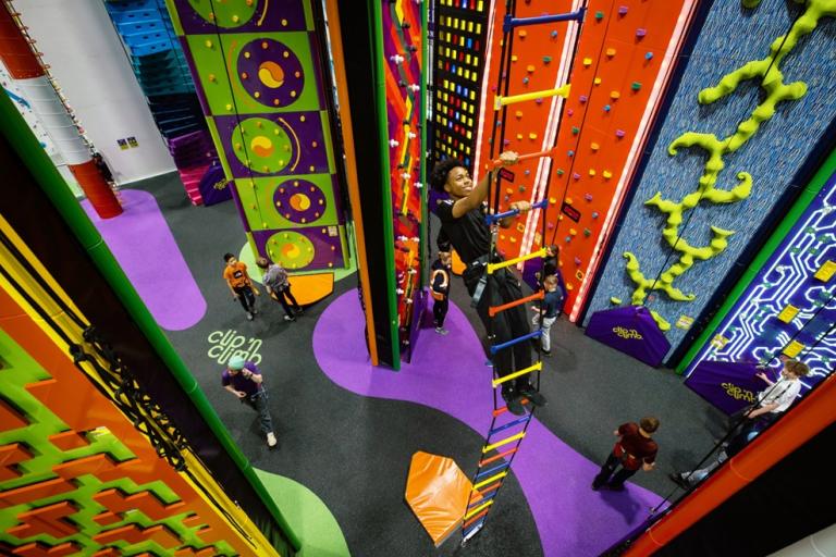 A colourful indoor climbing centre with various themed climbing walls, a hanging ladder, and several people climbing or waiting on the padded floor below.