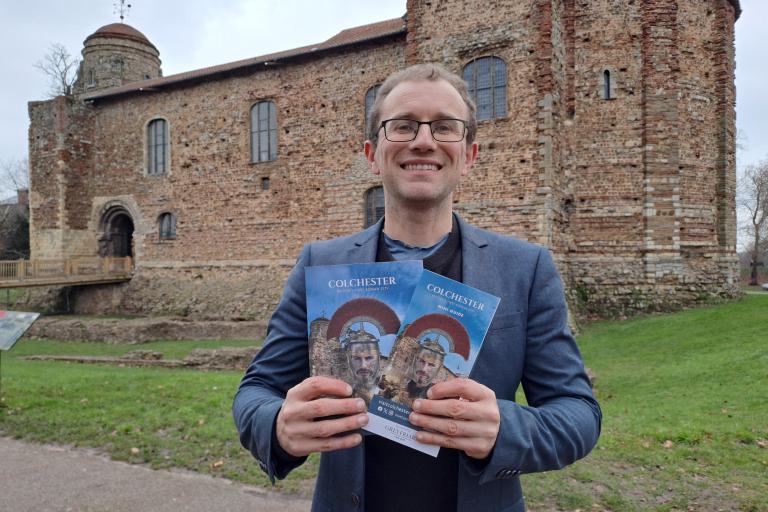 Cllr Lee Scordis holding Visit Colchester Guide in front of Colchester Castle