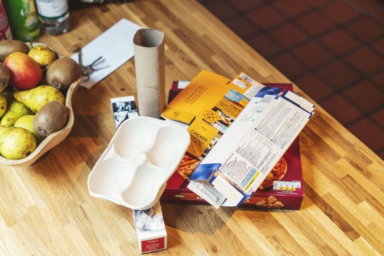 Recyclable cardboard and paper items laid out on a wooden kitchen counter beside a bowl of fruit.