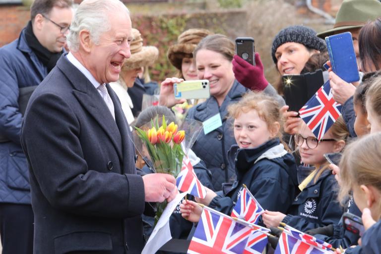 King Charles III holds a bouquet of yellow and red tulips while greeting a line of schoolchildren holding small Union Jack flags, with adults gathered behind them taking photos during an outdoor public visit.
