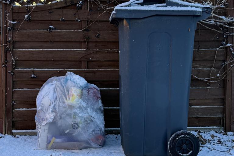 A clear plastic bag full of recycled paper and a black wheelie bin side by side in the snow
