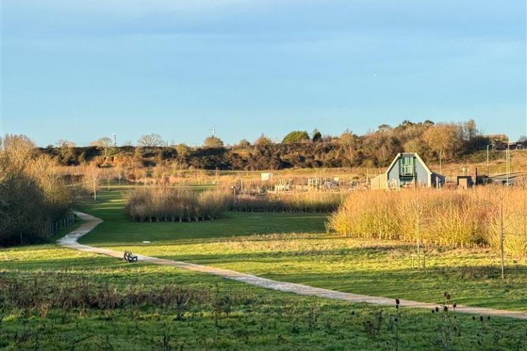 A wide, sunny landscape view of a country park with open grassy fields, a winding footpath, scattered trees and shrubs. A modern building sits to the right, with low hills and woodland in the background under a clear sky.