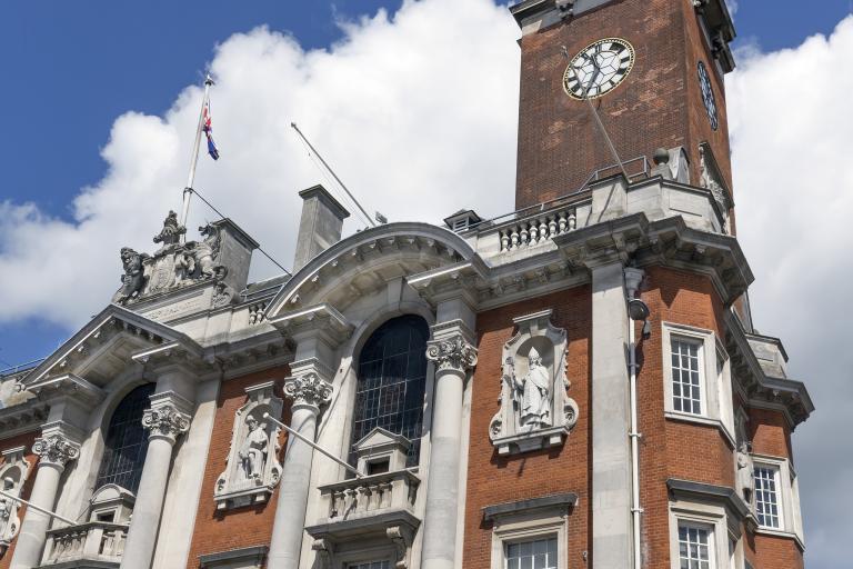 A tall historic brick building (Colchester Town Hall) with ornate stone detailing, arched windows, and classical statues set into the façade. A prominent clock tower rises above the structure, topped with decorative architectural features. The sky behind it is bright blue with scattered white clouds.
