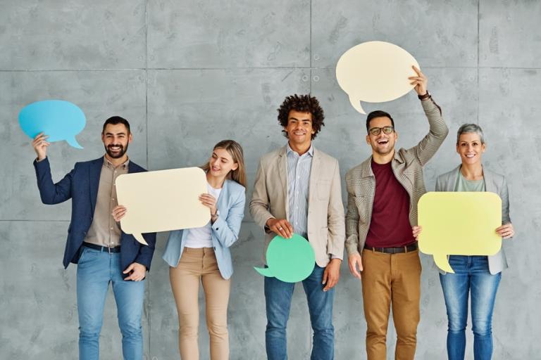 Five people standing against a gray textured wall, each holding large colorful speech bubble cutouts in various shapes. The speech bubbles are in shades of blue, yellow, green, and beige, suggesting themes of communication, conversation, or collaboration. The individuals are dressed in business-casual attire, and the overall setting conveys a modern, professional environment.