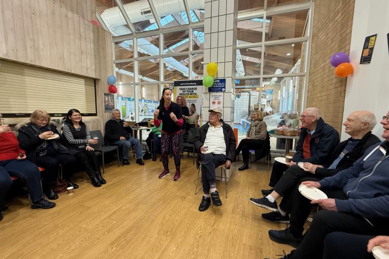 A group of people sit in a community space decorated with colourful balloons while a woman stands speaking in the centre of the room. Attendees are seated in a circle, holding refreshments, and the setting appears warm and social with large windows providing natural light.