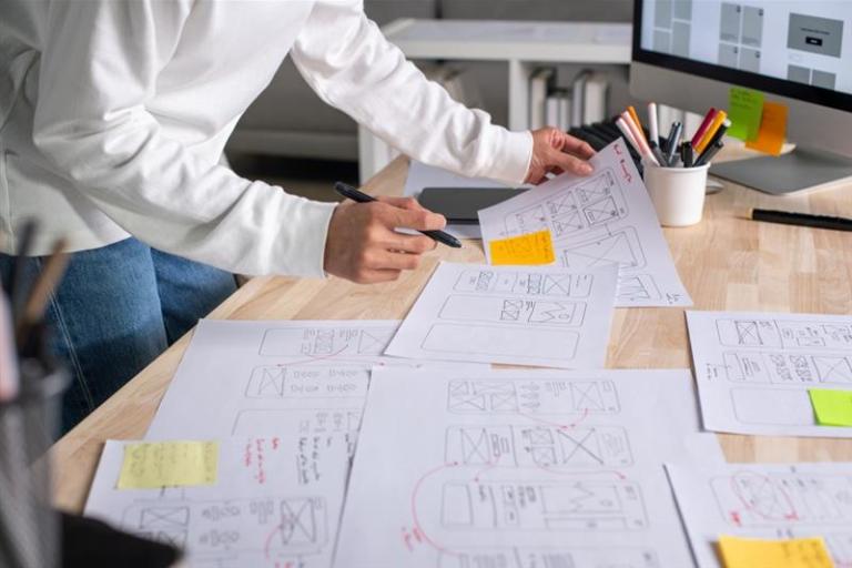 A person standing at a desk reviewing and marking UX wireframe sketches spread across the workspace, with a computer monitor and stationery in the background.