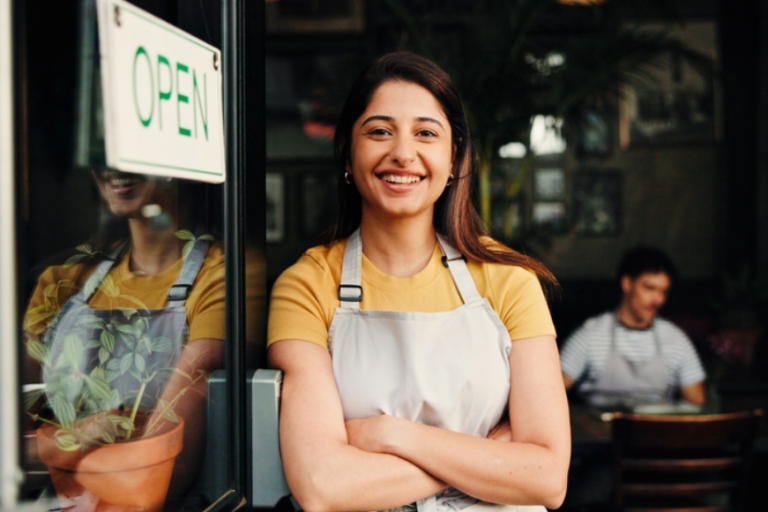 A photo of a person smiling in the door of a shop, there is a sign on the door that says 'open'