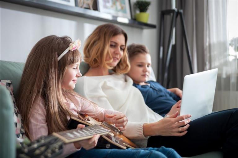 A scene of three individuals sitting together on a sofa in a living room. One person holds a laptop, while another sits nearby with a guitar resting across their lap. The room features soft natural light, wall décor on a shelf, and a floor lamp in the background.