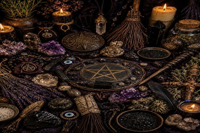 A flat lay of candles, crystals, herbs, pentacle symbols, charms and small ritual objects on a dark surface