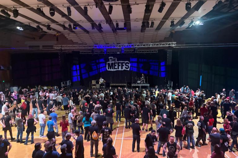 A large crowd stands inside Charter Hall facing a stage with “The Meffs” backdrop.