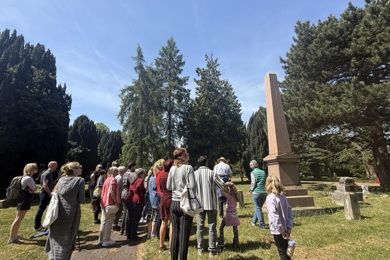 A group of people gathered around a tall obelisk monument among trees in Colchester Cemetery.