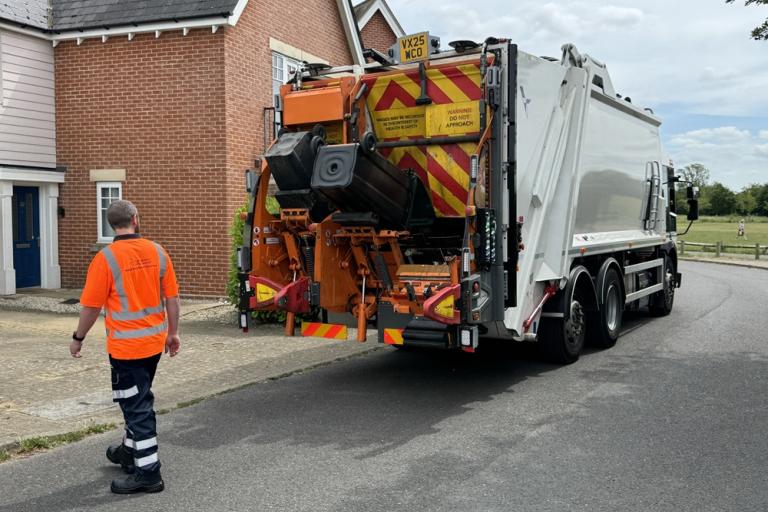 Refuse collection lorry collecting bins outside houses, with a worker in high-visibility clothing nearby.