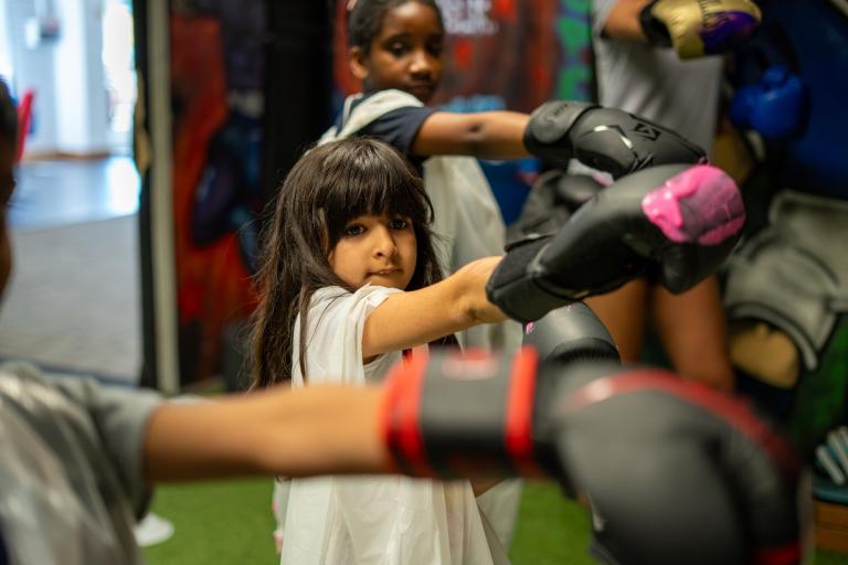 A group of children wearing boxing gloves practise punches on pads in a gym.