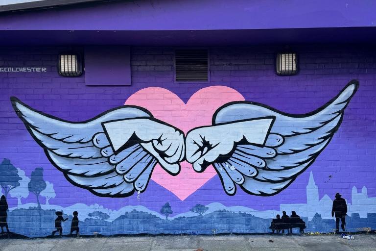 A large mural painted on a purple brick wall showing two white winged hands touching knuckles in front of a pink heart. Silhouettes of people, trees and a city skyline run along the bottom of the artwork, with a paved walkway in the foreground and a cloudy sky above.