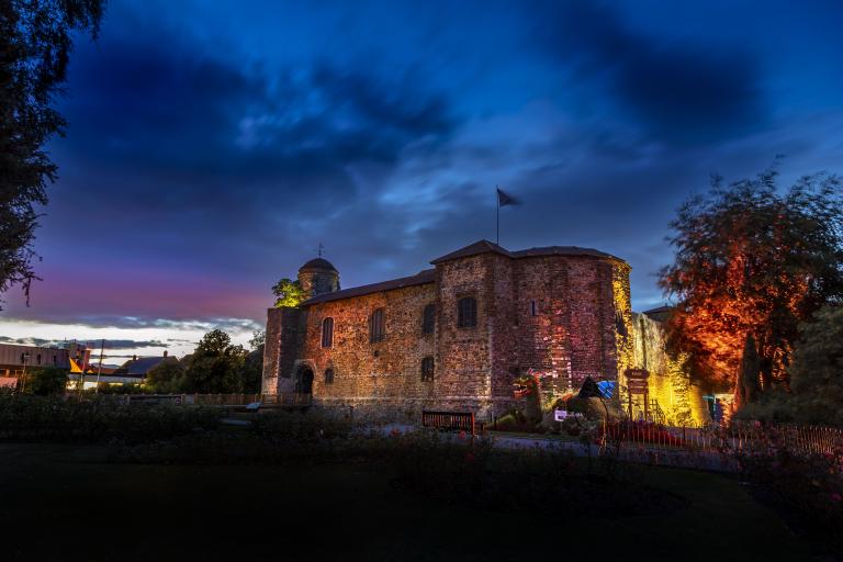 Colchester Castle illuminated at dusk beneath a dramatic blue and purple evening sky. The historic stone castle is lit with warm amber, pink and gold floodlights that highlight its textured walls and arched doorway. Trees frame the scene on both sides, with one brightly illuminated in orange and red light. A flag flies from the castle roof, and gardens, pathways, benches and visitor signage are visible in the foreground. 