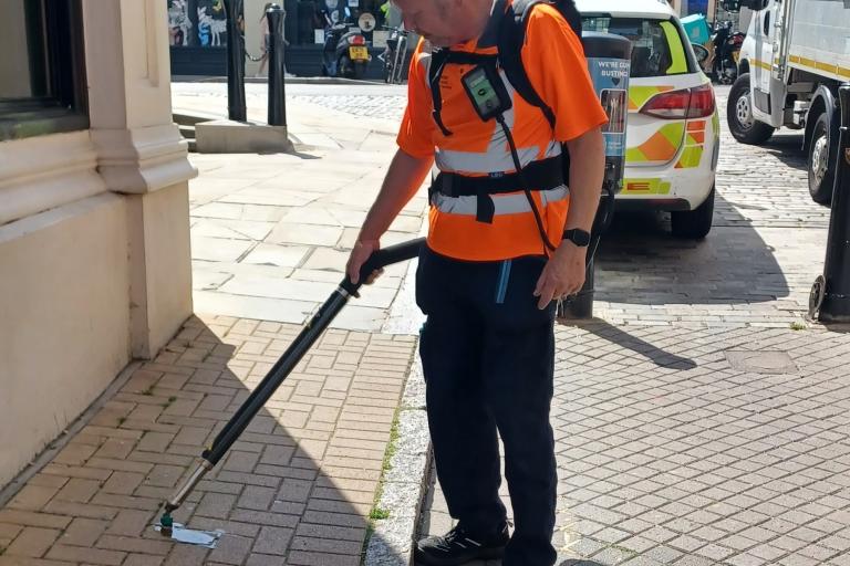 A person uses a long tool to remove chewing gum from a pavement beside a street.
