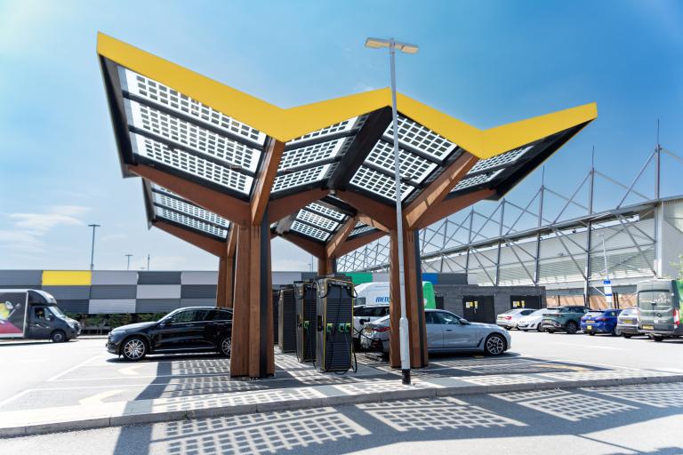 Solar-panel canopy sheltering electric vehicle charging points in a car park beside a large building.