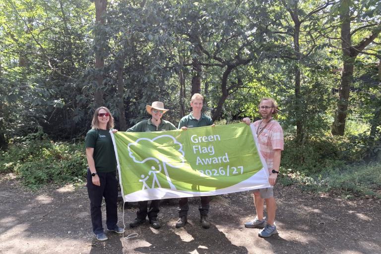 Four people hold a Green Flag Award 2026/27 banner in a wooded park.