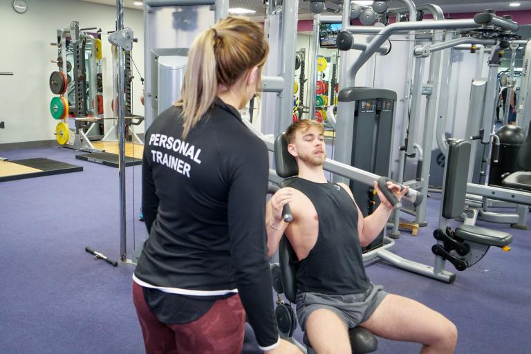 Person in 'Personal Trainer' top observing another using a chest press machine