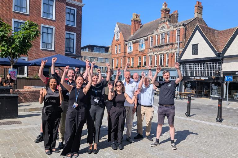 A group of around 12 people stand together in a paved town square with both arms raised in the air. The group is positioned in front of a row of historic red-brick and timber-fronted buildings under a clear blue sky. Outdoor café umbrellas and a small tree are visible on the left, while a building with signs reading “Other Monkey Brewing” appears in the background on the right. The scene is brightly lit by sunshine, with long shadows cast across the square.