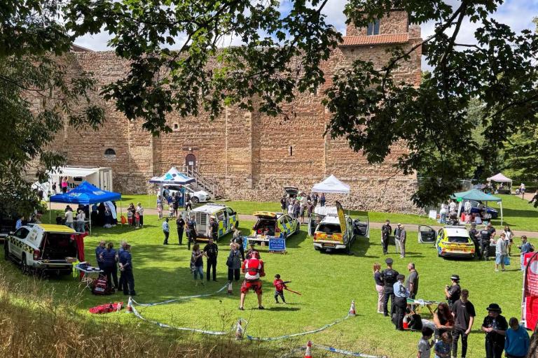 A community event on a grassy lawn beside a large historic stone building, with display tents, emergency service vehicles and groups of people.