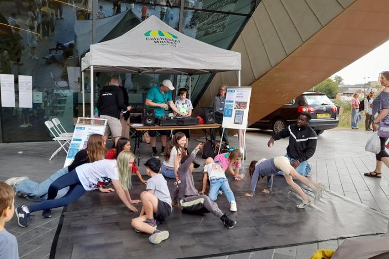 People on a dance mat beneath a market stall canopy outside a modern building