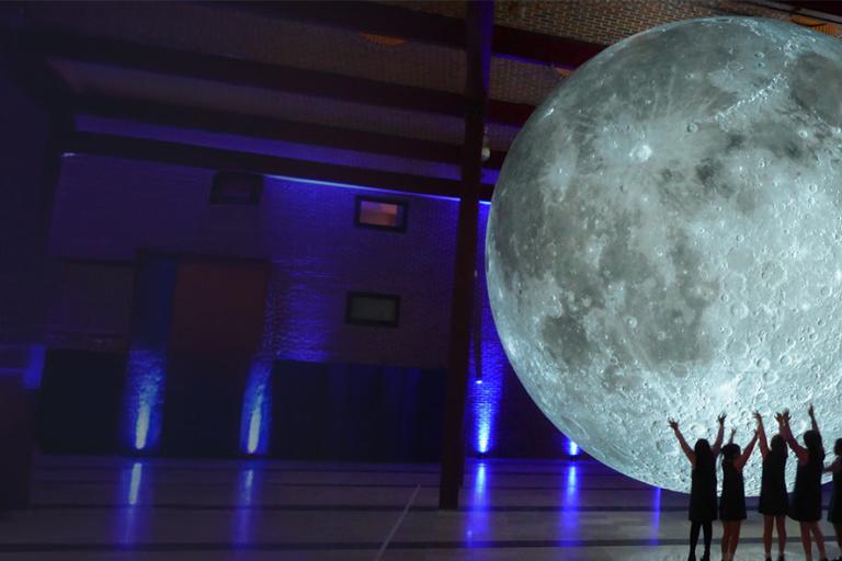 Large glowing moon in a hall and people standing beside it.