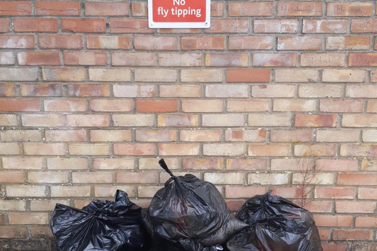 Three dumped black bags in front of a brick wall with a sign saying "No Fly Tipping"