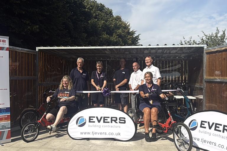 A group of people at a ribbon-cutting ceremony beside adapted cycles in a covered outdoor area.