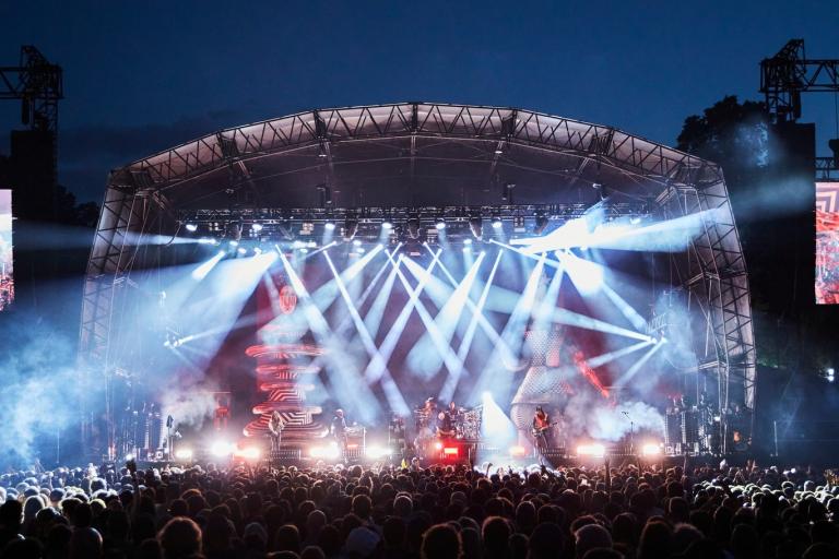 A large outdoor concert stage illuminated by bright lights, with a crowd gathered in front at night.