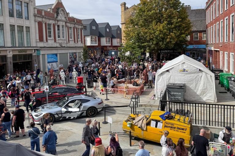 A busy city centre square with crowds, display vehicles, market stalls and surrounding buildings.