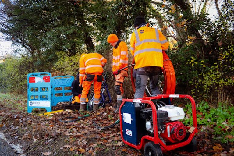 A group of people in high-visibility clothing working beside a roadside cable installation with equipment and orange ducting.