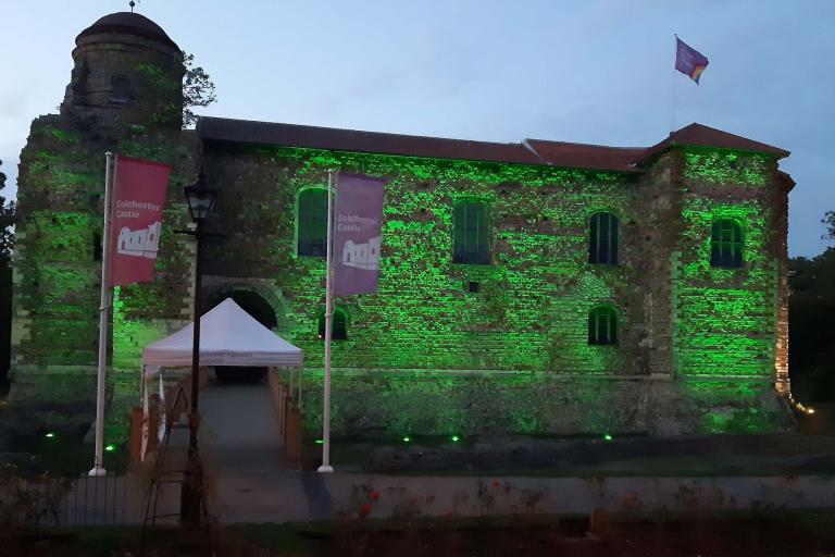 Colchester Castle at dusk, illuminated with green lighting across its historic stone walls. A pathway and bridge lead to the main entrance beneath a white canopy. Two flags reading "Colchester Castle" stand in front of the building, and a rainbow flag flies from a flagpole on the roof. Trees frame the castle on both sides beneath an overcast evening sky.