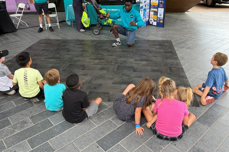 A person kneels on a mat demonstrating moves to seated children at an event.