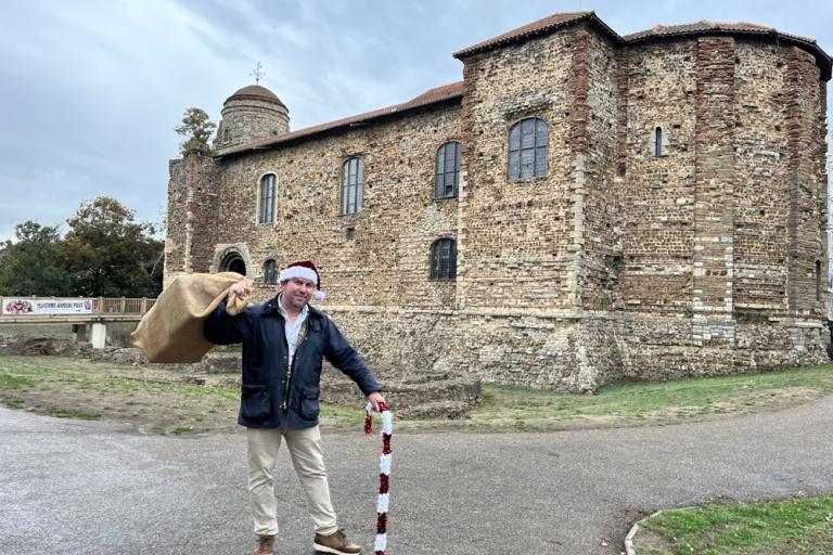 Cllr Darius Laws wearing a Christmas hat and carrying a sack of presents in front of Colchester Castle