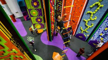 A colourful indoor climbing centre with various themed climbing walls, a hanging ladder, and several people climbing or waiting on the padded floor below.