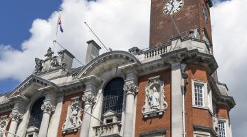 A tall historic brick building (Colchester Town Hall) with ornate stone detailing, arched windows, and classical statues set into the façade. A prominent clock tower rises above the structure, topped with decorative architectural features. The sky behind it is bright blue with scattered white clouds.