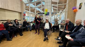 A group of people sit in a community space decorated with colourful balloons while a woman stands speaking in the centre of the room. Attendees are seated in a circle, holding refreshments, and the setting appears warm and social with large windows providing natural light.