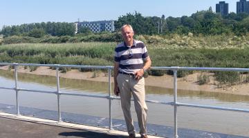 Cllr Sunnucks standing on a paved quayside path by a metal railing beside a tidal river, with marshland, trees and modern buildings in the background under a clear blue sky.