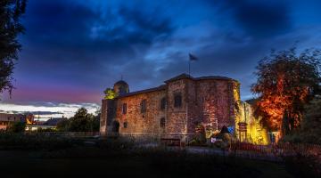 Colchester Castle illuminated at dusk beneath a dramatic blue and purple evening sky. The historic stone castle is lit with warm amber, pink and gold floodlights that highlight its textured walls and arched doorway. Trees frame the scene on both sides, with one brightly illuminated in orange and red light. A flag flies from the castle roof, and gardens, pathways, benches and visitor signage are visible in the foreground. 