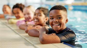 A group of children leaning on the edge of an indoor swimming pool in a line.