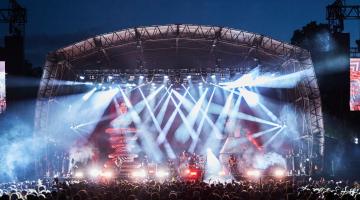 A large outdoor concert stage illuminated by bright lights, with a crowd gathered in front at night.