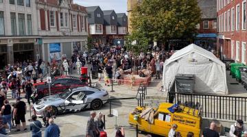A busy city centre square with crowds, display vehicles, market stalls and surrounding buildings.
