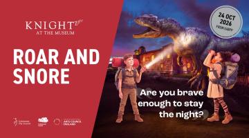 Two people with backpacks and torches stand in front of a dinosaur and museum at night.