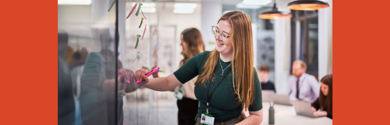A woman with long hair and glasses smiling and writing on a white board in a meeting room