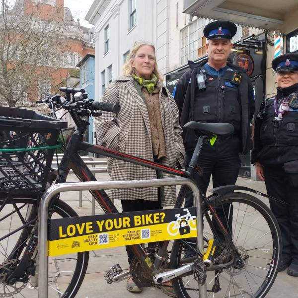 Three people stand beside a bicycle secured to a bike rack on Colchester High Street with a sign promoting secure bike parking.